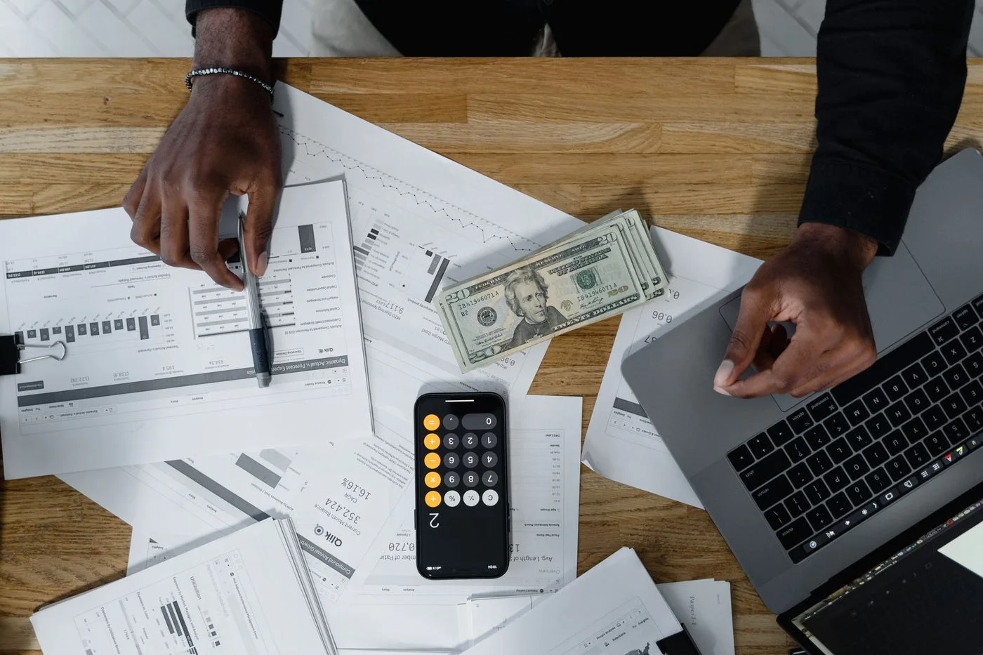 Hands calculate finances with papers, cash, and a laptop on a wooden desk.