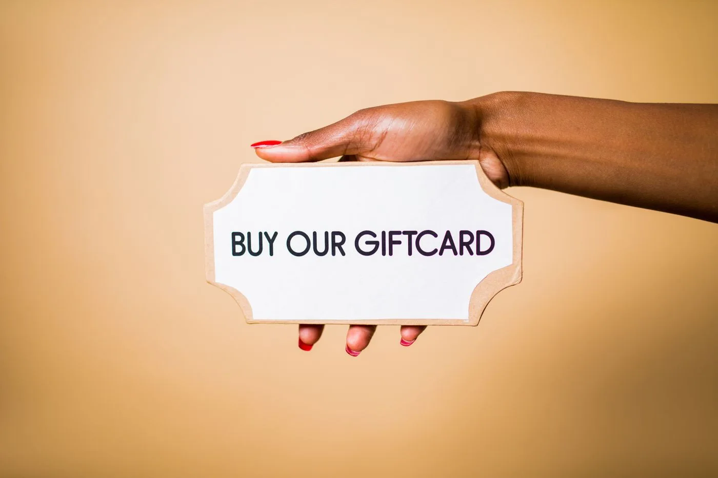 Close-up of a hand holding a 'Buy Our Giftcard' sign on a brown backdrop.