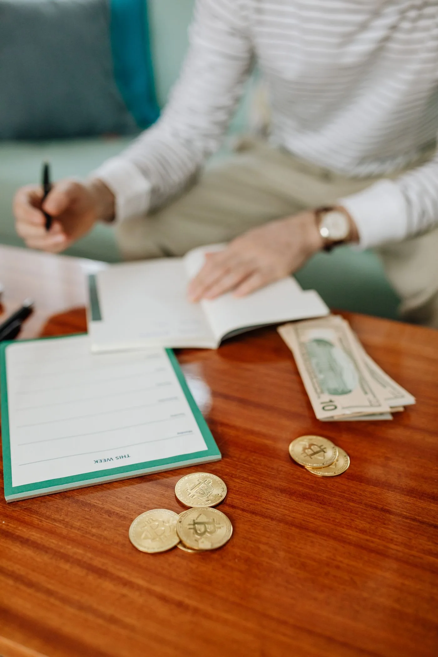 Person writing in a notebook with cash and coins on a wooden table, planning finances.