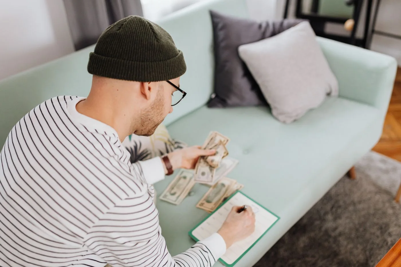 man counting money and writing notes while sitting indoors on a couch.