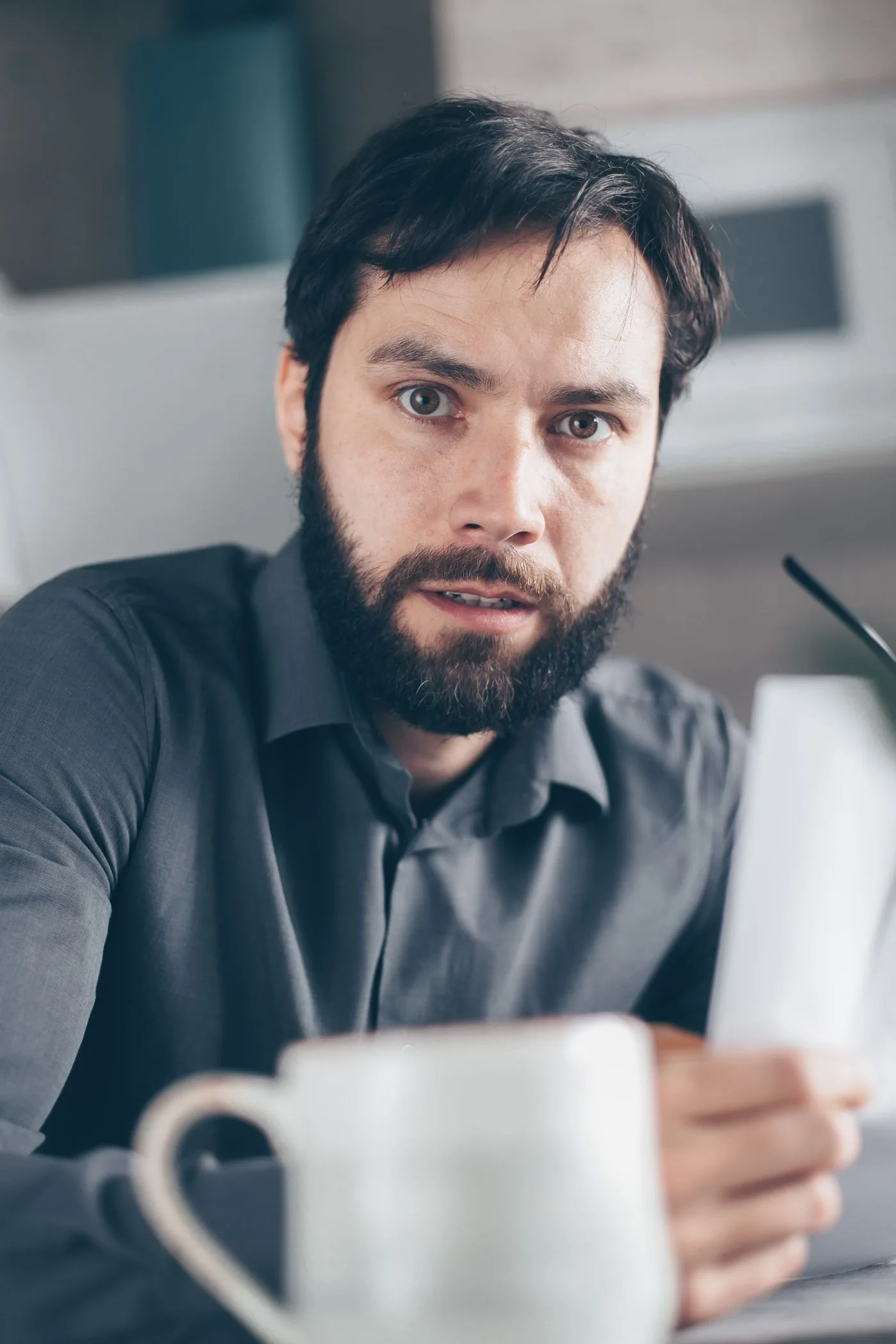 Worried man with beard reviewing financial paperwork indoors, holding cup.