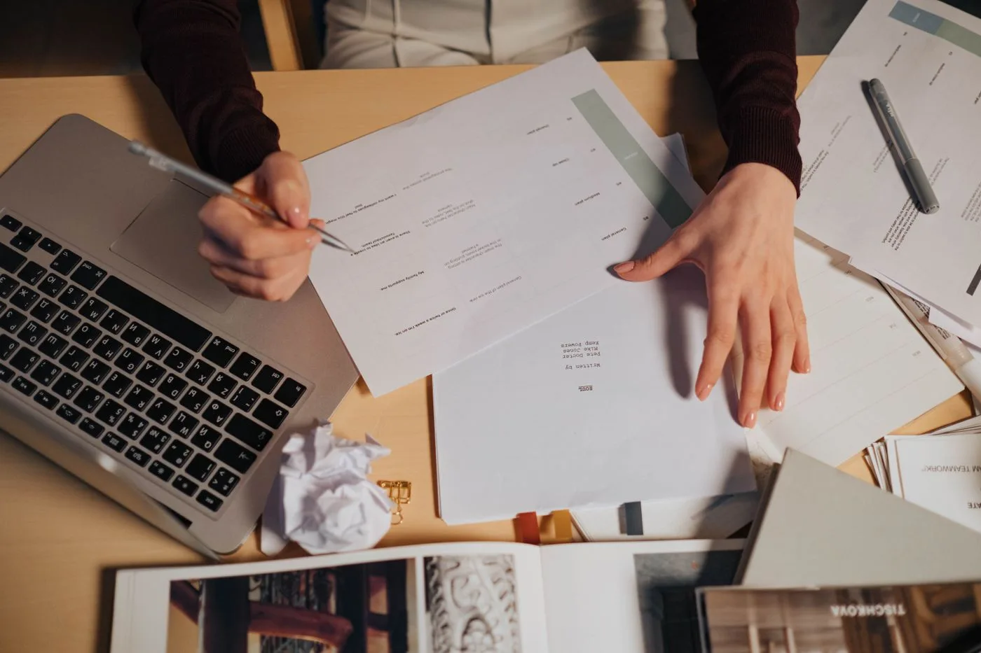 A person taking notes and working on a laptop surrounded by documents on a desk.