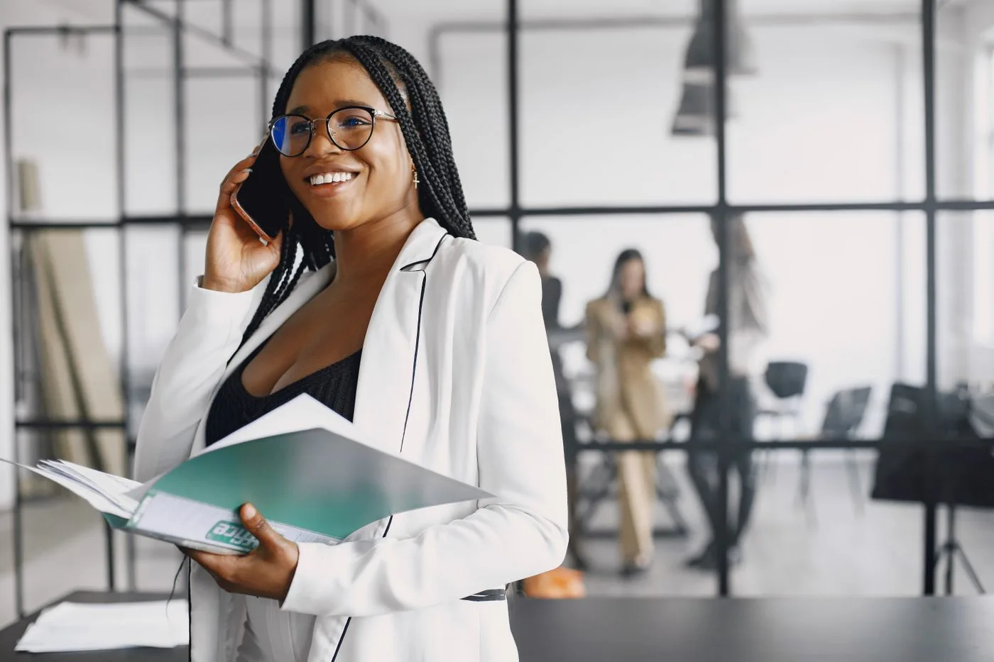 Confident black woman in a white blazer holding documents and talking on a phone in a stylish office.